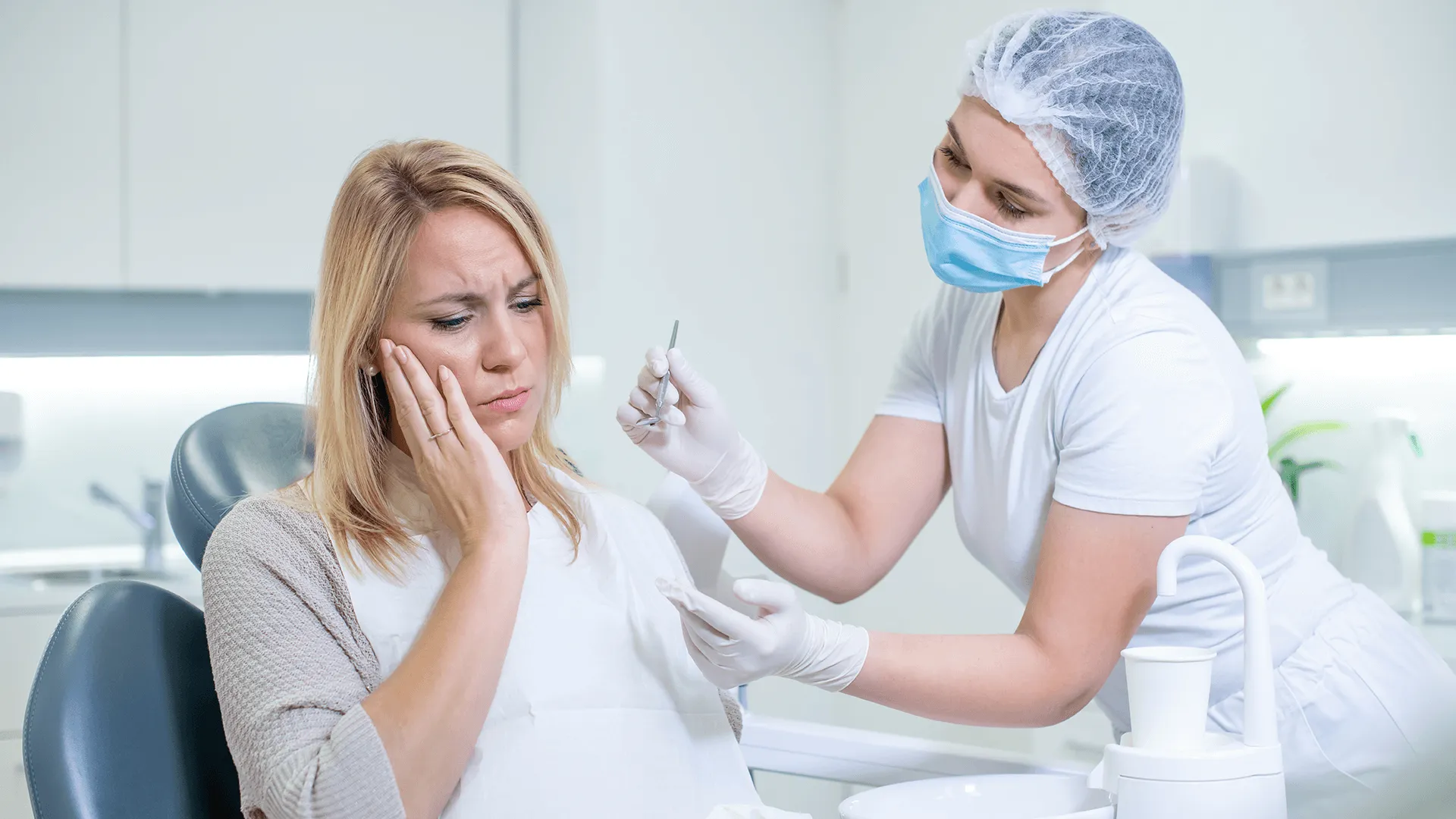 Middle shot of a Caucasian woman with a toothache sitting in the dental chair while a female dentist trying to examine her.