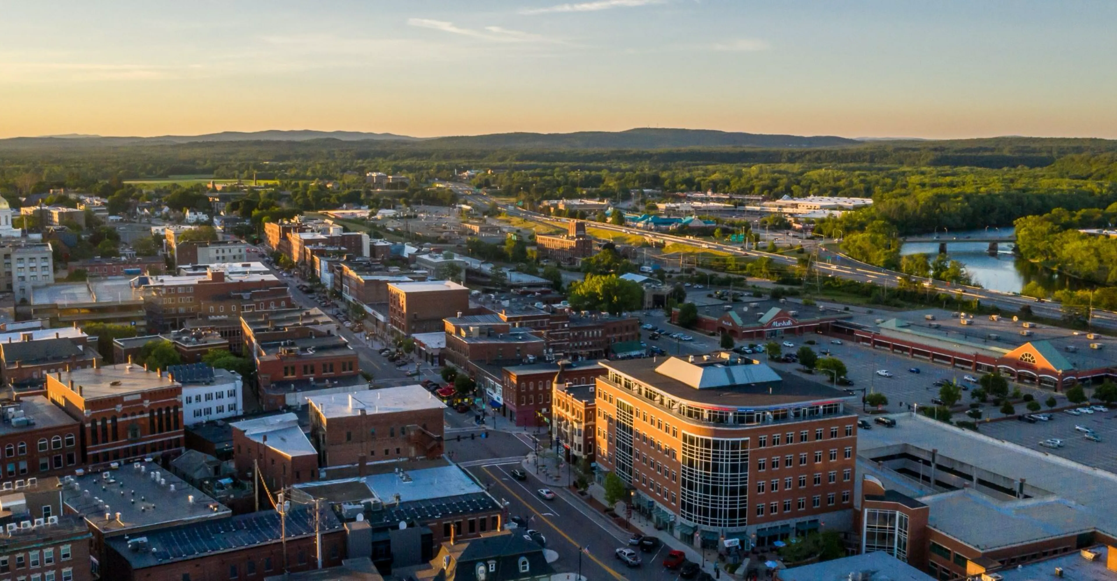 Aerial view of a cityscape at sunset, featuring buildings, roads, and greenery in the background.