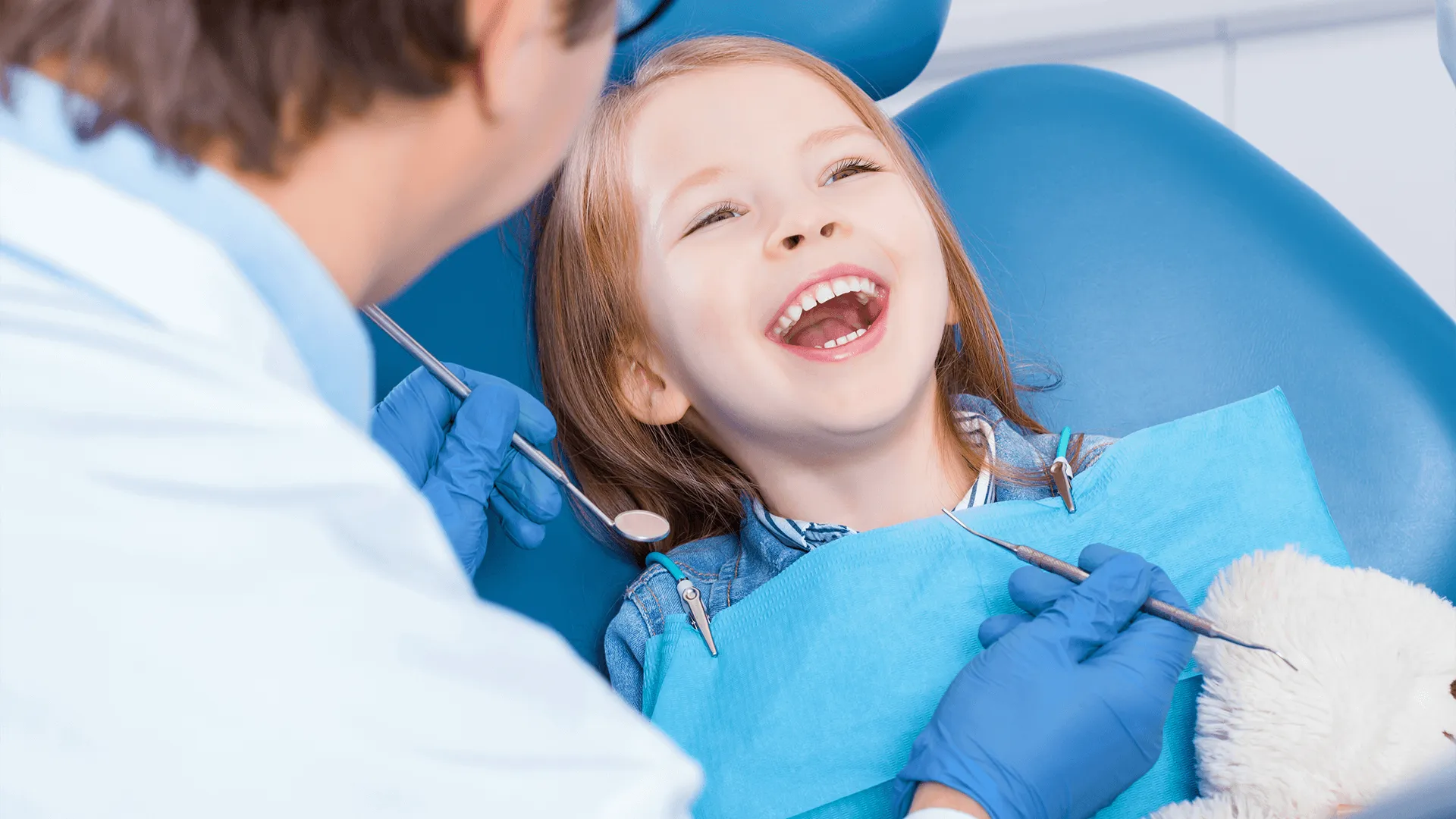 Little cute smiling girl is sitting in dental chair in clinic, office. Doctor is preparing for examination of child teeth with tools, amusing patient with bear toy